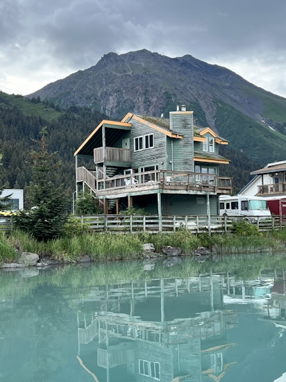 A wooden cabin with a rustic design is situated by a calm body of water, reflecting the building's structure on the surface. The cabin is surrounded by lush greenery, with a dense forest and towering mountain in the background. The sky is overcast, adding a serene and tranquil feel to the scene.