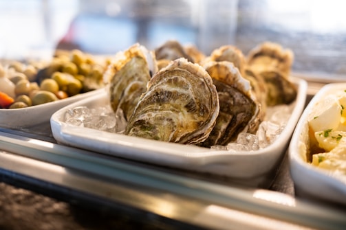 A close-up of a seafood display featuring oysters on a bed of ice in a white ceramic dish. The image also shows an assortment of olives in one dish and a partially visible dish containing a creamy potato salad. The lighting highlights the textures and natural colors of the seafood and side dishes.