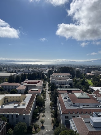 An aerial view captures a university campus with symmetrical pathways lined by large, academic buildings featuring red-tiled roofs and surrounded by green trees. In the background, a bay glistens under a partly cloudy sky.