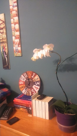 A wooden table features a collection of books, a colorful clock, a smartphone, and a television remote. A purple potted orchid plant with white flowers is placed on the right side. Two pieces of artwork hang on a blue wall above the table.