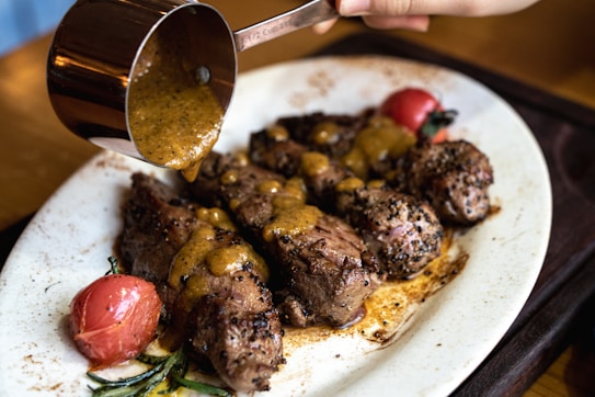 A plate of grilled steak pieces topped with a rich, golden sauce is being prepared. The sauce is being poured from a metal measuring cup onto the steaks. Cherry tomatoes and green herbs accompany the dish, adding a touch of color and freshness.