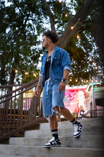 A man wearing a denim jacket over a black shirt, distressed denim shorts, and sneakers with high black socks stands on outdoor steps near a handrail. In the background, there are trees with hanging lights and an illuminated structure with colorful signage.