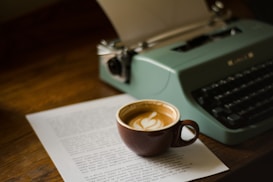 A vintage typewriter sits on a wooden table beside a cup of coffee with latte art. Underneath the coffee cup, there is a printed document with text visible.