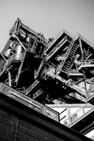 A monochrome industrial structure with intricate metal frameworks and staircases, angled upwards capturing both the geometric complexity and the massive scale of the construction.
