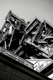 A monochrome industrial structure with intricate metal frameworks and staircases, angled upwards capturing both the geometric complexity and the massive scale of the construction.