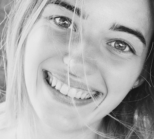 A close-up black and white photograph of a person's smiling face, with hair partially covering the eyes. The smile is broad and genuine, revealing teeth, and the skin texture is visible due to the lighting and monochrome effect.