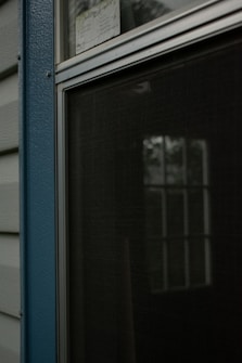 A closed window with a metal frame and a fly screen, reflecting some greenery outside. It is positioned beside textured blue paneling and light-colored siding.