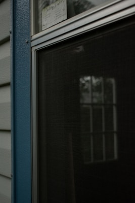 A closed window with a metal frame and a fly screen, reflecting some greenery outside. It is positioned beside textured blue paneling and light-colored siding.