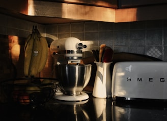 A cozy kitchen counter with a retro toaster, a white stand mixer, and a basket of bananas. Soft, warm lighting casts shadows and highlights various textures, including tiled backsplash and stainless steel surfaces. Utensils are kept in a ceramic holder, adding to the tidy appearance.