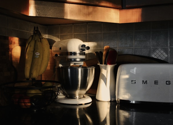A cozy kitchen counter with a retro toaster, a white stand mixer, and a basket of bananas. Soft, warm lighting casts shadows and highlights various textures, including tiled backsplash and stainless steel surfaces. Utensils are kept in a ceramic holder, adding to the tidy appearance.