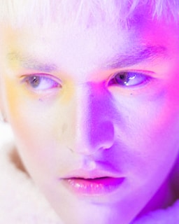 A close-up of a person's face, highlighting their eyes and lips. The lighting casts a blend of vibrant purple, pink, and yellow hues across the skin, creating an ethereal and artistic effect.