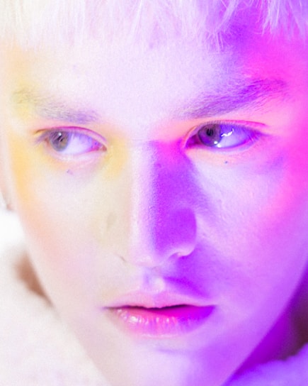 A close-up of a person's face, highlighting their eyes and lips. The lighting casts a blend of vibrant purple, pink, and yellow hues across the skin, creating an ethereal and artistic effect.