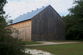A large, rustic barn with wooden walls and a metal roof stands in a grassy area surrounded by trees. The barn has tall, narrow doors and a weathered appearance, with a mix of aged gray and natural wood tones. A gravel path leads up to the barn, and the sky above is partly cloudy.