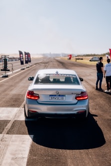 A white BMW car is on a race track with a license plate reading 'BMW5E38'. The car is positioned at the starting line. On the right side, two people are standing and talking. The track features racing flags and a timing light structure. The scene is set on a clear day with a bright blue sky.