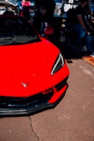 A red sports car with sleek lines and a modern design is parked on a paved surface. The front part of the car shows a prominent emblem, and its headlights are sharply angled. In the background, several people are gathered, some standing and others seated, with some vehicles parked nearby.