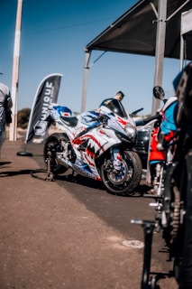 A sports motorcycle parked on a concrete surface under a large canopy. The motorcycle features a custom paint job with red, white, and blue colors, displaying graphics of teeth and eyes. Nearby, a flag with the word 'UNIQUE' is displayed, and several people are present, with one person standing close to the motorcycle. Other motorcycles and equipment are visible in the background.