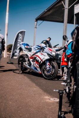 A sports motorcycle parked on a concrete surface under a large canopy. The motorcycle features a custom paint job with red, white, and blue colors, displaying graphics of teeth and eyes. Nearby, a flag with the word 'UNIQUE' is displayed, and several people are present, with one person standing close to the motorcycle. Other motorcycles and equipment are visible in the background.