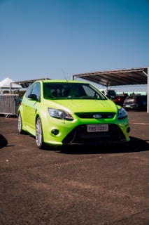 A vibrant green sporty hatchback car is parked on an asphalt surface under a clear blue sky. The car is positioned head-on, with a visible license plate and distinctive design features highlighting its performance nature. In the background, several structures and cars are partially visible, contributing to an open and spacious atmosphere.