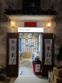 An inviting, rustic shop entrance with wooden doors propped open, revealing a colorful display of products inside. Traditional lanterns hang on either side of the entrance, casting a warm glow. The exterior wall has a textured, aged appearance.