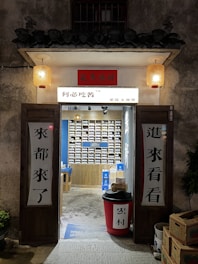 An inviting, rustic shop entrance with wooden doors propped open, revealing a colorful display of products inside. Traditional lanterns hang on either side of the entrance, casting a warm glow. The exterior wall has a textured, aged appearance.
