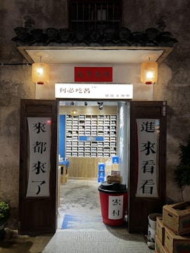 An inviting, rustic shop entrance with wooden doors propped open, revealing a colorful display of products inside. Traditional lanterns hang on either side of the entrance, casting a warm glow. The exterior wall has a textured, aged appearance.