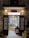 An inviting, rustic shop entrance with wooden doors propped open, revealing a colorful display of products inside. Traditional lanterns hang on either side of the entrance, casting a warm glow. The exterior wall has a textured, aged appearance.