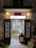 An inviting, rustic shop entrance with wooden doors propped open, revealing a colorful display of products inside. Traditional lanterns hang on either side of the entrance, casting a warm glow. The exterior wall has a textured, aged appearance.