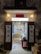 An inviting, rustic shop entrance with wooden doors propped open, revealing a colorful display of products inside. Traditional lanterns hang on either side of the entrance, casting a warm glow. The exterior wall has a textured, aged appearance.