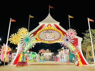 Brightly lit entrance to a themed amusement park or carnival at night. The entrance structure is shaped like a circus tent with red and white stripes, adorned with an illuminated sign that reads 'Circus Land.' Colorful flower-shaped light decorations flank the entrance, and there are various whimsical decorations including a clown statue and other playful elements. Flags are mounted on top of tall poles, and additional lights and signs are visible further inside.