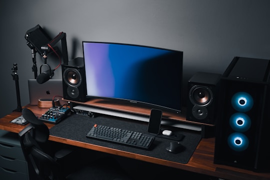 A modern gaming and streaming setup with a curved monitor placed centrally on a wooden desk. Surrounding the monitor are powerful speakers and a high-end PC tower with glowing fans. Various peripherals, including a mechanical keyboard, ergonomic mouse, and audio mixer, are neatly arranged. Overhead, a professional microphone and stand are ready for use, possibly for gaming commentary or podcasting.