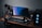A modern gaming and streaming setup with a curved monitor placed centrally on a wooden desk. Surrounding the monitor are powerful speakers and a high-end PC tower with glowing fans. Various peripherals, including a mechanical keyboard, ergonomic mouse, and audio mixer, are neatly arranged. Overhead, a professional microphone and stand are ready for use, possibly for gaming commentary or podcasting.
