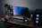 A modern gaming and streaming setup with a curved monitor placed centrally on a wooden desk. Surrounding the monitor are powerful speakers and a high-end PC tower with glowing fans. Various peripherals, including a mechanical keyboard, ergonomic mouse, and audio mixer, are neatly arranged. Overhead, a professional microphone and stand are ready for use, possibly for gaming commentary or podcasting.