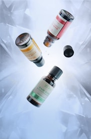 Three bottles of hair tonic are suspended in mid-air, with one open cap and a droplet falling from one of the bottles. The bottles have labels in different colors: yellow, red, and green. The background is a light, icy blue with an abstract, crystalline pattern.