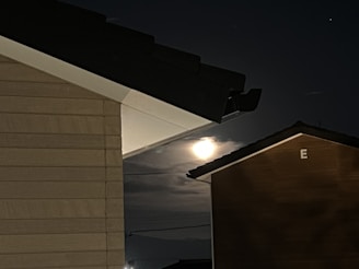 A night scene capturing a bright moon partially obscured by clouds. The image is framed by the rooftops and walls of two buildings, with one building labeled with the letter 'E'. The buildings have dark roofs that contrast against the dimly lit sky.
