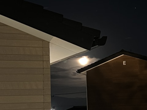 A night scene capturing a bright moon partially obscured by clouds. The image is framed by the rooftops and walls of two buildings, with one building labeled with the letter 'E'. The buildings have dark roofs that contrast against the dimly lit sky.