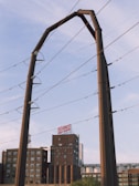 A large industrial structure, possibly for electrical wiring, is set against a partly cloudy sky. In the background, there is a multi-story brick building with a sign on top and various other industrial elements.
