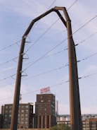 A large industrial structure, possibly for electrical wiring, is set against a partly cloudy sky. In the background, there is a multi-story brick building with a sign on top and various other industrial elements.