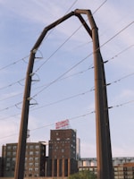 A large industrial structure, possibly for electrical wiring, is set against a partly cloudy sky. In the background, there is a multi-story brick building with a sign on top and various other industrial elements.