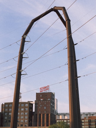 A large industrial structure, possibly for electrical wiring, is set against a partly cloudy sky. In the background, there is a multi-story brick building with a sign on top and various other industrial elements.