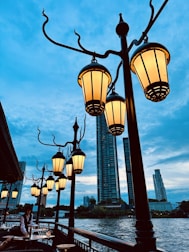 A series of ornate street lamps with warm glowing lights line a riverside walkway, set against a backdrop of tall city buildings and a clear blue sky. A person, possibly a waiter, is attending to a table along the outdoor seating area.
