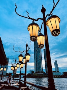 A series of ornate street lamps with warm glowing lights line a riverside walkway, set against a backdrop of tall city buildings and a clear blue sky. A person, possibly a waiter, is attending to a table along the outdoor seating area.