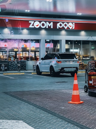 A modern gas station scene featuring a white Range Rover with black rims parked in front of a convenience store named Zoom. The store has large glass windows displaying shelves stocked with various products. A bright orange traffic cone is placed in the foreground next to a fire extinguisher on a cart. The area is well-lit with overhead lights, and the pavement is made of patterned tiles.