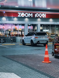 A modern gas station scene featuring a white Range Rover with black rims parked in front of a convenience store named Zoom. The store has large glass windows displaying shelves stocked with various products. A bright orange traffic cone is placed in the foreground next to a fire extinguisher on a cart. The area is well-lit with overhead lights, and the pavement is made of patterned tiles.