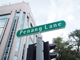A street sign labeled 'Penang Lane' is prominently displayed in the foreground. In the background, there's a tall, modern building with large reflective windows. To the side, the tops of trees are visible under a partly cloudy sky.