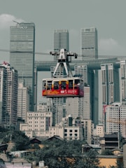 A red cable car is suspended in mid-air, traveling across a modern cityscape filled with tall skyscrapers and densely packed buildings. The background features high-rise towers with a mix of architectural styles, including a distinctive building with a horizontal passage connecting its vertical sections. The lower part of the image includes smaller residential structures with greenery visible below.
