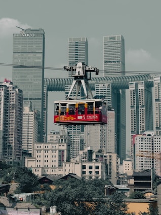A red cable car is suspended in mid-air, traveling across a modern cityscape filled with tall skyscrapers and densely packed buildings. The background features high-rise towers with a mix of architectural styles, including a distinctive building with a horizontal passage connecting its vertical sections. The lower part of the image includes smaller residential structures with greenery visible below.