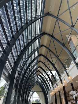 The image captures the interior of a modern, well-lit architectural space with a high, vault-like ceiling supported by large, curved metal beams. The ceiling features a combination of glass and wooden panels. On the right, there are multiple hanging light fixtures and a second-floor balcony area with glass railings. The design gives the space an open and airy feel.