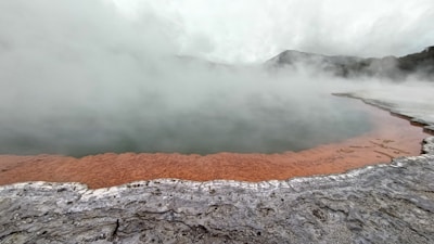 A geothermal landscape with a steaming body of water surrounded by mineral-rich formations. The edges are tinged with an orange hue, contrasting against the gray, rocky surface. Mist rises above the water, shrouding the background and partially obscuring distant hills.