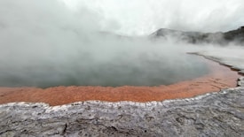 A geothermal landscape with a steaming body of water surrounded by mineral-rich formations. The edges are tinged with an orange hue, contrasting against the gray, rocky surface. Mist rises above the water, shrouding the background and partially obscuring distant hills.