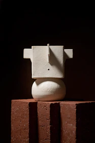A minimalist sculpture made of recycled metal pieces, displayed on a wooden pedestal in natural light.